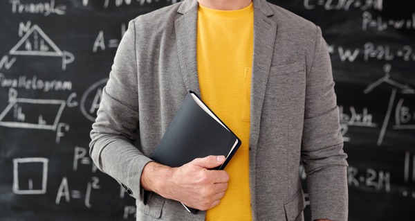Profesor delante de una pizarra sujetando un cuaderno.